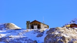 A cabin rests on a snowy mountain with a clear blue sky. The structure is rustic, surrounded by rocky terrain with patches of snow. Another cabin is visible in the distance, highlighting the remote and serene location.