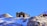 A cabin rests on a snowy mountain with a clear blue sky. The structure is rustic, surrounded by rocky terrain with patches of snow. Another cabin is visible in the distance, highlighting the remote and serene location.