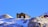 A cabin rests on a snowy mountain with a clear blue sky. The structure is rustic, surrounded by rocky terrain with patches of snow. Another cabin is visible in the distance, highlighting the remote and serene location.
