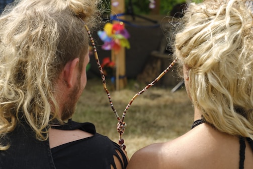 Two people are sitting closely with their backs turned, both having long, light-colored curly hair. They are connected by a strand of beads. A colorful pinwheel is visible in the background, along with some greenery.