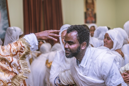 A group of people dressed in white garments are gathered, with one individual at the center being sprinkled with water by another person in ornate attire. The mood is focused and solemn with an indoor setting.