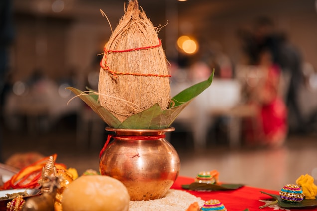 A decorated bronze pot contains a coconut wrapped in red thread and resting on green leaves. Surrounding the main setup are colorful decorative items, a small idol, and grains placed on a red cloth.