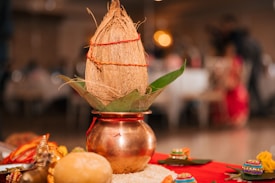 A decorated bronze pot contains a coconut wrapped in red thread and resting on green leaves. Surrounding the main setup are colorful decorative items, a small idol, and grains placed on a red cloth.