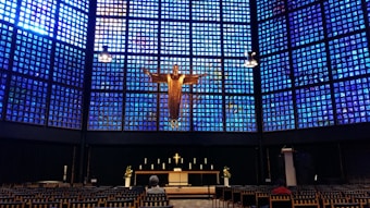 A church interior features a striking blue glass wall behind the altar, with a large golden figure of Christ prominently displayed. The altar is adorned with candles and floral arrangements, and the foreground includes rows of chairs, with a person seated among them.
