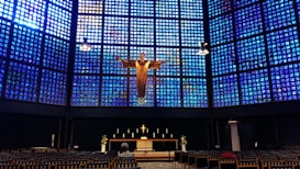 A church interior features a striking blue glass wall behind the altar, with a large golden figure of Christ prominently displayed. The altar is adorned with candles and floral arrangements, and the foreground includes rows of chairs, with a person seated among them.