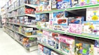 A brightly lit toy store aisle with shelves stocked with various boxed toys, including model cars, helicopters, building blocks, and plush toys. The shelves are organized with items of different shapes and colors, and the packaging features colorful designs and cartoon characters.