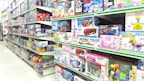 A brightly lit toy store aisle with shelves stocked with various boxed toys, including model cars, helicopters, building blocks, and plush toys. The shelves are organized with items of different shapes and colors, and the packaging features colorful designs and cartoon characters.