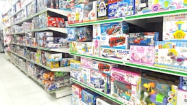 A brightly lit toy store aisle with shelves stocked with various boxed toys, including model cars, helicopters, building blocks, and plush toys. The shelves are organized with items of different shapes and colors, and the packaging features colorful designs and cartoon characters.