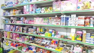 A store shelf filled with various colorful stationery items and art supplies, including pens, pencils, notebooks, and paint sets. The shelves are well-organized, displaying products in various vibrant packages. Some toy items and educational kits are also visible.