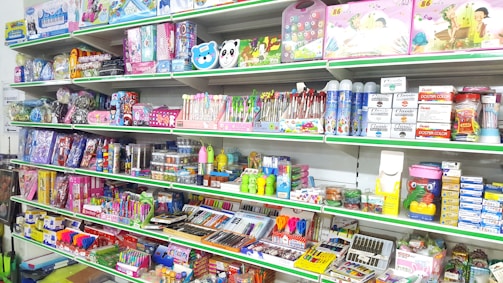 A store shelf filled with various colorful stationery items and art supplies, including pens, pencils, notebooks, and paint sets. The shelves are well-organized, displaying products in various vibrant packages. Some toy items and educational kits are also visible.