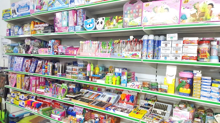 A store shelf filled with various colorful stationery items and art supplies, including pens, pencils, notebooks, and paint sets. The shelves are well-organized, displaying products in various vibrant packages. Some toy items and educational kits are also visible.