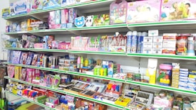 A store shelf filled with various colorful stationery items and art supplies, including pens, pencils, notebooks, and paint sets. The shelves are well-organized, displaying products in various vibrant packages. Some toy items and educational kits are also visible.