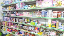 A store shelf filled with various colorful stationery items and art supplies, including pens, pencils, notebooks, and paint sets. The shelves are well-organized, displaying products in various vibrant packages. Some toy items and educational kits are also visible.