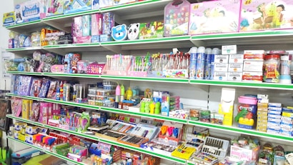 A store shelf filled with various colorful stationery items and art supplies, including pens, pencils, notebooks, and paint sets. The shelves are well-organized, displaying products in various vibrant packages. Some toy items and educational kits are also visible.