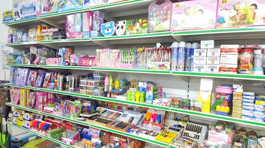 A store shelf filled with various colorful stationery items and art supplies, including pens, pencils, notebooks, and paint sets. The shelves are well-organized, displaying products in various vibrant packages. Some toy items and educational kits are also visible.