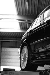 A black car with a shiny exterior is positioned inside an industrial setting. The focus is on the car's rear and wheel, with large garage doors and steel beams visible in the background. The image is captured in black and white, emphasizing the metallic and sleek surfaces.