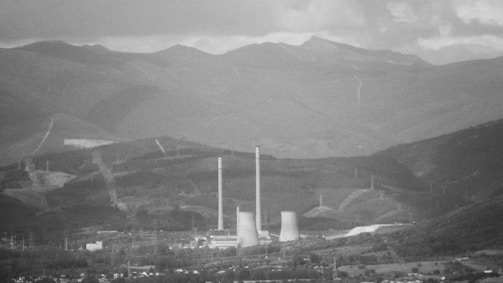 A grayscale landscape featuring a power plant with two tall smokestacks and cooling towers surrounded by mountainous terrain. The distant mountains are enveloped in a layer of mist, and the sky is overcast with a thick layer of clouds.