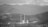 A grayscale landscape featuring a power plant with two tall smokestacks and cooling towers surrounded by mountainous terrain. The distant mountains are enveloped in a layer of mist, and the sky is overcast with a thick layer of clouds.