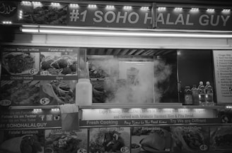 A black and white photo of a busy food truck displaying various menu items such as chicken kebab, falafel over rice, and gyro. The name of the vendor '#1 Soho Halal Guy' is prominently displayed. There is steam rising from the cooking area, indicating active cooking. Bottles of beverages are visible on a counter.
