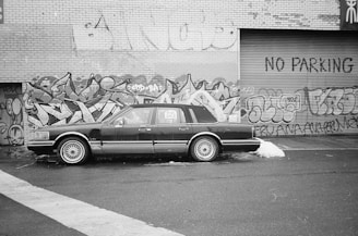 A black and white photograph of a classic car parked in front of a graffiti-covered wall. The vehicle has a 'For Sale' sign in one window. The wall is made of brick and has various colorful graffiti designs along with the words 'NO PARKING' painted on the metal shutter.