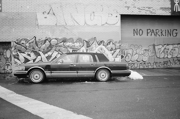 A black and white photograph of a classic car parked in front of a graffiti-covered wall. The vehicle has a 'For Sale' sign in one window. The wall is made of brick and has various colorful graffiti designs along with the words 'NO PARKING' painted on the metal shutter.