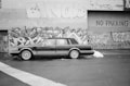 A black and white photograph of a classic car parked in front of a graffiti-covered wall. The vehicle has a 'For Sale' sign in one window. The wall is made of brick and has various colorful graffiti designs along with the words 'NO PARKING' painted on the metal shutter.