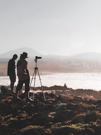Silhouettes of two people stand near a tripod with a camera setup on rocky terrain overlooking a body of water. The background features misty hills and a hazy sky, creating a serene and contemplative scene.