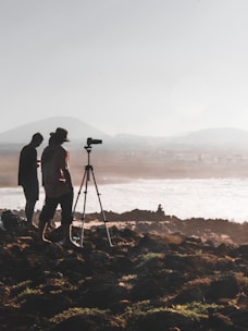 Silhouettes of two people stand near a tripod with a camera setup on rocky terrain overlooking a body of water. The background features misty hills and a hazy sky, creating a serene and contemplative scene.