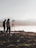 Silhouettes of two people stand near a tripod with a camera setup on rocky terrain overlooking a body of water. The background features misty hills and a hazy sky, creating a serene and contemplative scene.