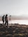 Silhouettes of two people stand near a tripod with a camera setup on rocky terrain overlooking a body of water. The background features misty hills and a hazy sky, creating a serene and contemplative scene.