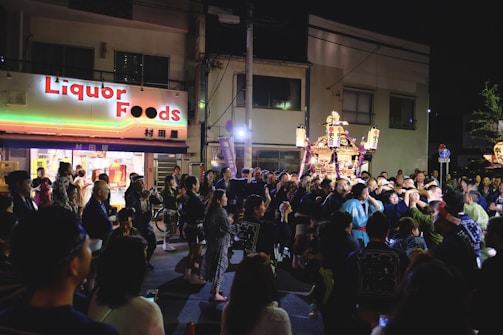 A vibrant night scene at a festival with a large group of people gathered around a decorated portable shrine. Many participants are wearing traditional clothing, such as happi coats. The shrine is ornately adorned and lit, creating a festive atmosphere. A store with a bright sign reading 'Liquor Foods' is visible in the background, along with various banners and signs.