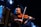 A musician plays the violin with a concentrated expression while standing on stage. The scene is dimly lit with spotlights creating a moody, atmospheric effect. In the foreground, a second instrument, possibly an accordion, is partially visible.