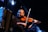 A musician plays the violin with a concentrated expression while standing on stage. The scene is dimly lit with spotlights creating a moody, atmospheric effect. In the foreground, a second instrument, possibly an accordion, is partially visible.