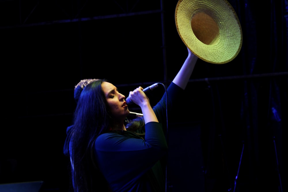 woman in blue long sleeved shirt holding up brown straw hat white talking in microphone