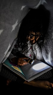 A child is reading a book under a blanket with a flashlight. The dim lighting and the cozy atmosphere suggest a secretive and intimate moment. The blanket has a star pattern, and the book is opened to a page with illustrations.