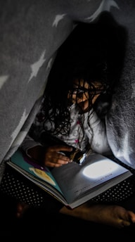 A child is reading a book under a blanket with a flashlight. The dim lighting and the cozy atmosphere suggest a secretive and intimate moment. The blanket has a star pattern, and the book is opened to a page with illustrations.