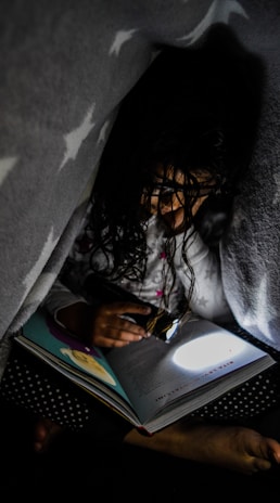 A child is reading a book under a blanket with a flashlight. The dim lighting and the cozy atmosphere suggest a secretive and intimate moment. The blanket has a star pattern, and the book is opened to a page with illustrations.