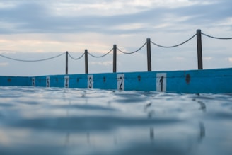 An outdoor swimming pool with lanes separated by numbered markers along the edge, with a chain fence in the background and the sky showing a mix of clouds and light blue hues.