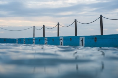 An outdoor swimming pool with lanes separated by numbered markers along the edge, with a chain fence in the background and the sky showing a mix of clouds and light blue hues.