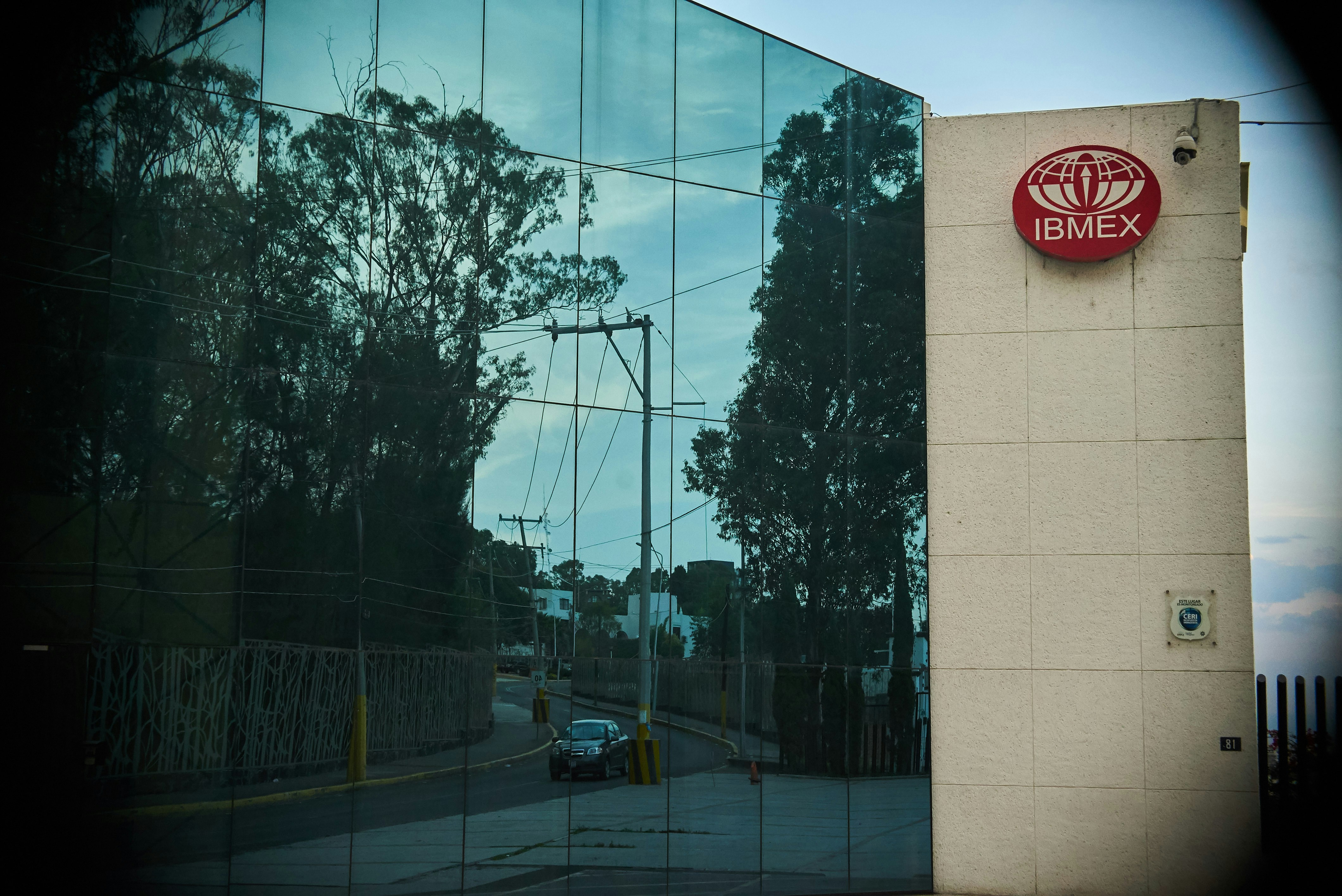 A modern building features a reflective glass facade displaying the reflection of trees and a clear sky. A red sign with the logo and text 'IBMEX' is mounted on a beige wall section. A road with a parked car and power lines is visible in the reflections.