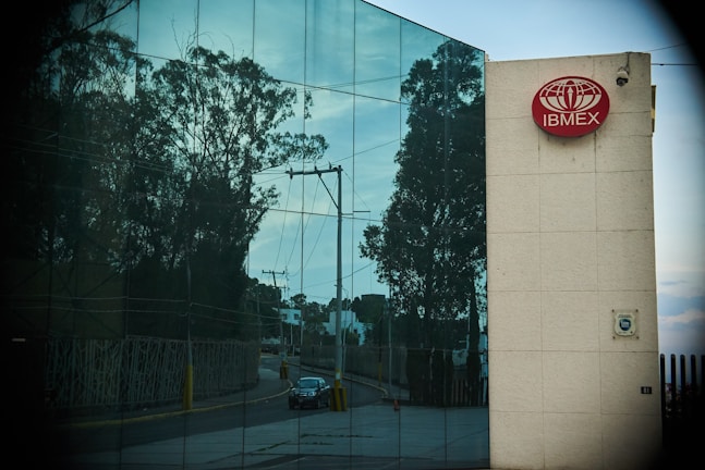 A modern building features a reflective glass facade displaying the reflection of trees and a clear sky. A red sign with the logo and text 'IBMEX' is mounted on a beige wall section. A road with a parked car and power lines is visible in the reflections.