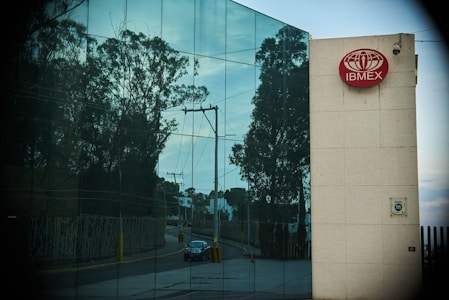 A modern building features a reflective glass facade displaying the reflection of trees and a clear sky. A red sign with the logo and text 'IBMEX' is mounted on a beige wall section. A road with a parked car and power lines is visible in the reflections.