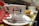 A white coffee cup with the logo of Manteigaria printed on it, featuring a handshake illustration. The cup is placed on a white saucer with a shiny metal teaspoon. A red cloth or napkin is folded next to the cup. In the blurred background, there is a pastry on a plate, suggesting a cafe setting.