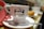 A white coffee cup with the logo of Manteigaria printed on it, featuring a handshake illustration. The cup is placed on a white saucer with a shiny metal teaspoon. A red cloth or napkin is folded next to the cup. In the blurred background, there is a pastry on a plate, suggesting a cafe setting.