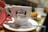 A white coffee cup with the logo of Manteigaria printed on it, featuring a handshake illustration. The cup is placed on a white saucer with a shiny metal teaspoon. A red cloth or napkin is folded next to the cup. In the blurred background, there is a pastry on a plate, suggesting a cafe setting.