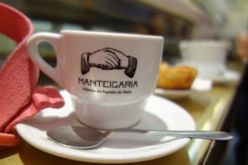 A white coffee cup with the logo of Manteigaria printed on it, featuring a handshake illustration. The cup is placed on a white saucer with a shiny metal teaspoon. A red cloth or napkin is folded next to the cup. In the blurred background, there is a pastry on a plate, suggesting a cafe setting.