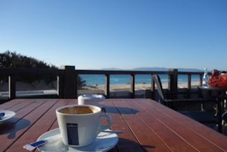 A cup of coffee sits on a wooden table overlooking a scenic view of a beach and ocean. The setting includes clear blue skies and a few scattered tables and chairs. A distant shoreline and low hills are visible on the horizon.