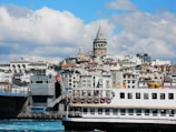 A bustling cityscape featuring a large ferry crossing turquoise waters in the foreground, with a backdrop of historic buildings and a prominent tower. The architecture is a mix of modern and classic styles, and a Turkish flag is visible. Fluffy clouds add depth to the blue sky.