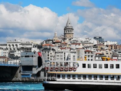 A bustling cityscape featuring a large ferry crossing turquoise waters in the foreground, with a backdrop of historic buildings and a prominent tower. The architecture is a mix of modern and classic styles, and a Turkish flag is visible. Fluffy clouds add depth to the blue sky.