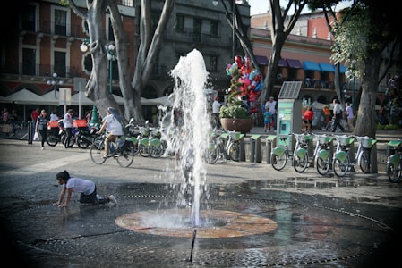 A public square features a water fountain at the center with a child playing near it. Bicycles are parked at a bike station, and several people are walking or cycling around. Trees and buildings line the area, with colorful balloons visible in the background.